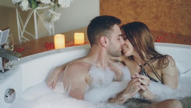 Young attractive lovers in bathing suits are hugging and kissing in jacuzzi with foam in day spa. Romantic relationship, passion, relaxation and wellness concept.