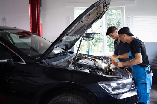 Manager checks tasts on tablet computer and explains an Engine breakdown to an male mechanic. Car service employees inspect car's engine bay with a LED lamp. Modern clean workshop.
