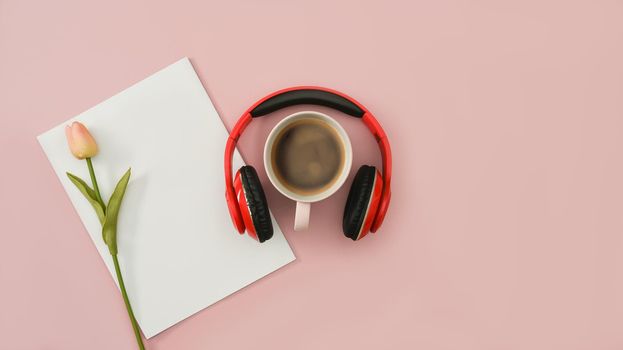 Red wireless headphones, coffee cup, blank paper and tulip on pink background. Top view, flat lay with copy space.