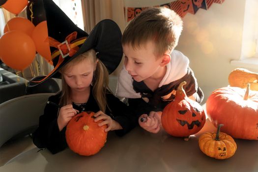 Halloween party at home. A little girl in big hat and witch costume and a boy in a skeleton costume are drawing pumpkins. The concept of kind, home holiday for the whole family. Children's creativity