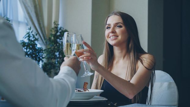 Attractive girlfriend and boyfriend in fance clothing are clinking glasses, drinking champagne and talking on romantic date in classy restaurant. Romance, love and fine dining concept.