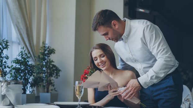 Beautiful woman is waiting for her boyfriend in nice restaurant, he is coming and giving her flowers, kissing her. Happy girl is smelling flowers and smiling with pleasure.