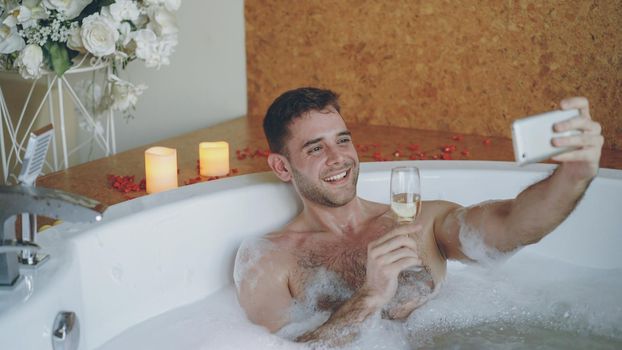 Handsome young man is taking selfie with champagne glass using smartphone in hot tub with foam in modern spa salon. He is smiling and posing looking at camera.