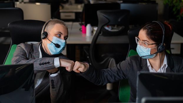 Two business women in masks are giving a high five while sitting at one desk in the office.