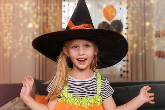 Happy little girl, blonde smiles and laughs. The child is dressed in a witch costume, a large black hat with a pointed end and a striped T-shirt. Home family for a party, Halloween holiday