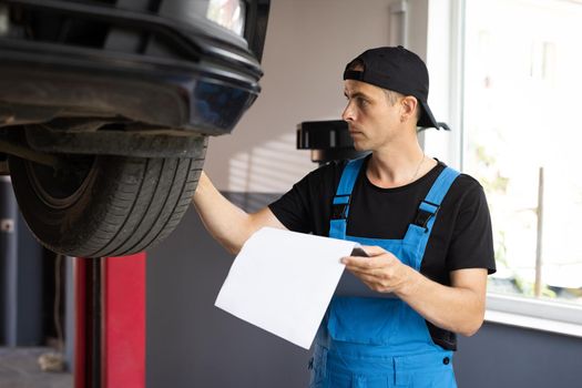 Car service employe inspect car. Mechanic inspects the car undercarriage way and makes a note on his inspection sheet. Automobile service, car mechanic. Modern workshop