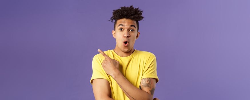 Portrait of surprised, excited young man showing cool new thing, pointing upper left corner, gasping, folding lips, stare camera impressed say wow, standing purple background.