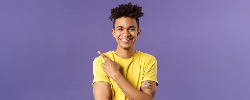 Close-up portrait of enthusiastic, happy young hipster male with dreads, beaming smile and pointing finger upper left corner, present cool product, introduce something really good, purple background.