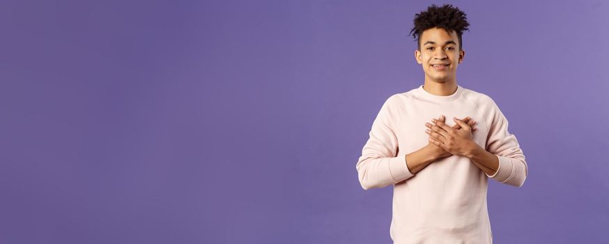 Portrait of romantic handsome hispanic man with dreads, hold hands pressed to heart and smiling pleased, see heartwarming nice gesture, receive lovely gift, being touched, purple background.