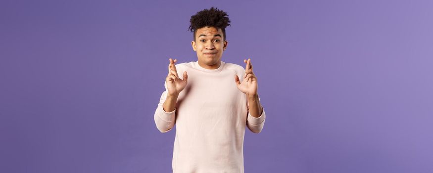 Portrait of young hopeful handsome man with dreads ancitipating something good happen, cross fingers good luck, smiling with joy and optimism, having faith, praying over purple background.