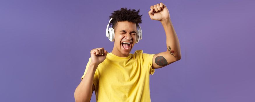 Close-up portrait of cheerful, happy young dancing guy lift hand up singing along, close eyes and smiling upbeat as listening awesome song in headphones, enjoying music, purple background.