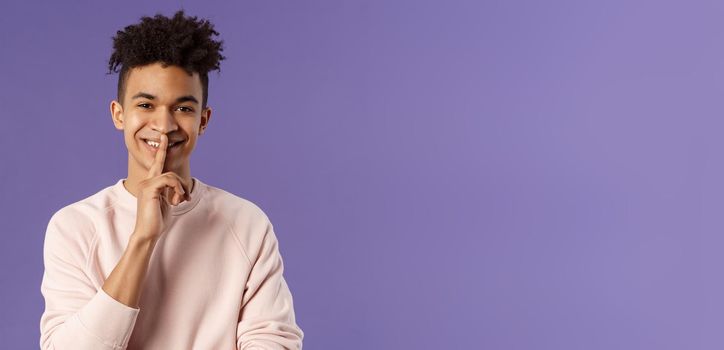 Close-up portrait of attractive smiling young man hiding secret, asking keep silent or quiet to do surprise, show shush gesture, place index finger to mouth, standing purple background.