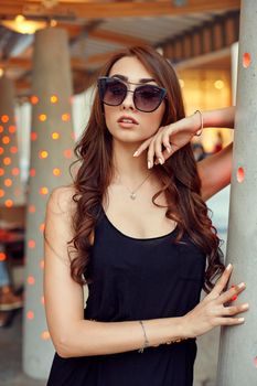 Close-up portrait of young elegant brunette in black dress and sunglasses, touching her face. Fashion street shot. Woman walking on the street, wearing trendy outfit, travel.