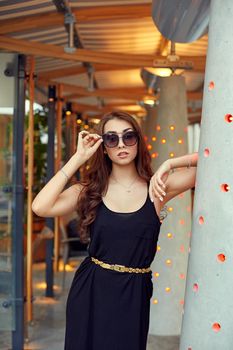 Close-up portrait of young elegant brunette in black dress and sunglasses. Fashion street shot. Woman walking on the street, wearing trendy outfit, travel.