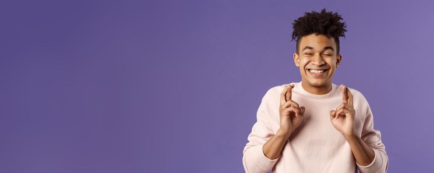 Close-up portrait of happy young hopeful guy anticipating something good happen, cross fingers good luck, close eyes and smiling awaiting miracle, praying for dream come true, purple background.