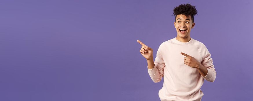 Portrait of upbeat, good-looking hispanic man with dreads, smiling amused, looking and pointing upper left corner, advertisement of product or company banner, copy space, purple background.