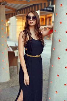 Close-up portrait of young elegant brunette in black dress and sunglasses. Fashion street shot. Woman walking on the street, wearing trendy outfit, travel.