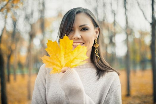 Young smiling woman in a gray sweater with an autumn yellow maple leaf in her hand. Autumn mood.