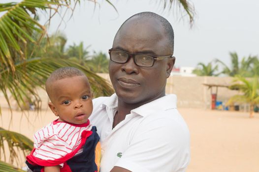 a man standing with his baby near a coconut tree and looking at the camera.