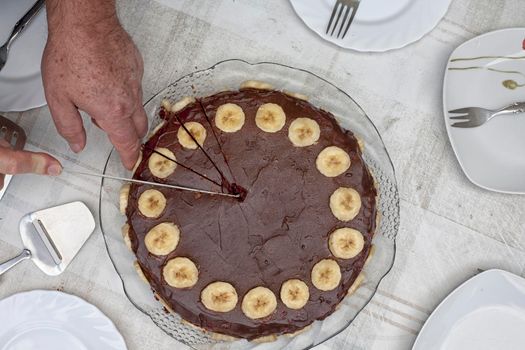 Flat lay of a chocholate cake. With slices of babanas along the edges. A compound of brown and yellow. You can see the hand and the knife cutting the cake.