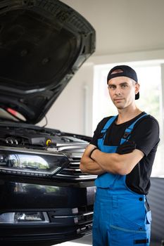 Portrait shot of caucasian man in blue uniform and hat standing in big auto garage, looking to camera and crossing hands. Pretty male mechanic in maintaining service.