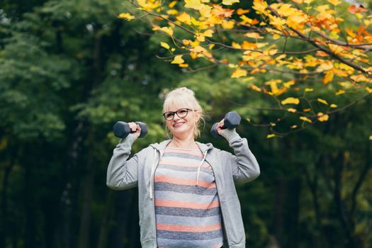 Senior woman standing and doing sports exercises with dumbbells in her hands in the park on the grass, exercising, leading a healthy active lifestyle sitting on the grass