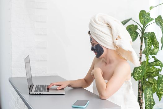 Young woman with towel on head and black facial clay mask sitting at home and using laptop computer . Staying at home, quarantine, distance learning, online education, technology, modern lifestyle.