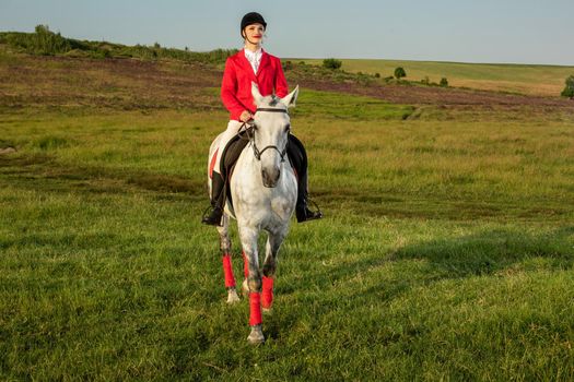 Young woman rider, wearing red redingote and white breeches, with her horse in evening sunset light. Outdoor photography in lifestyle mood