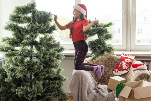 little girl with long hair decorates a green tree. Celebrating a Christmas party. The child decorates the Christmas tree at home.