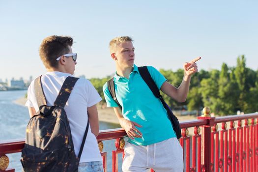 Outdoor portrait of two friends boys teenagers 15, 16 years old, talking laughing. Guys standing on bridge over river on sunny summer day. Youth, friendship, communication