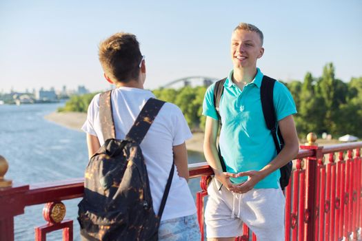 Outdoor portrait of two friends teenagers boys 15, 16 years old, laughing, talking in sunny day, standing on bridge over the river. Urban lifestyle, teens, friendship, communication