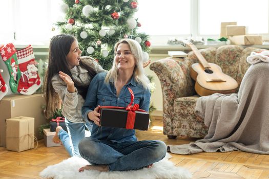Merry Christmas and Happy Holidays. Cheerful mother and her cute daughter girl exchanging gifts. Mom and little child having fun near tree indoors. Loving family with presents in room.