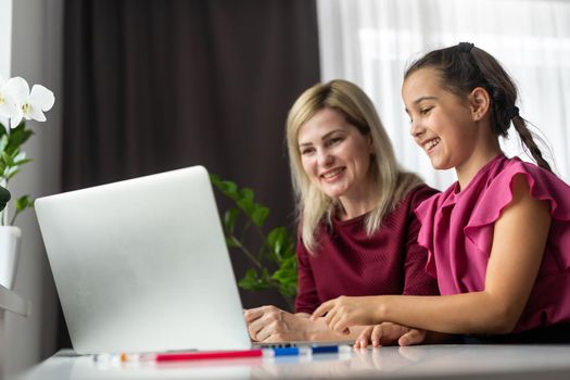 Pleased mother and small daughter sitting at table and school supplies and laptop and doing home task together in apartment.