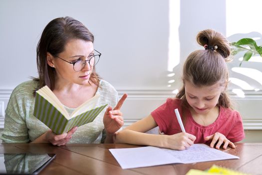 Mother and daughter child study together at home, sitting at table. Woman with book, girl writes in notebook. Distance learning, parent helping child primary school student