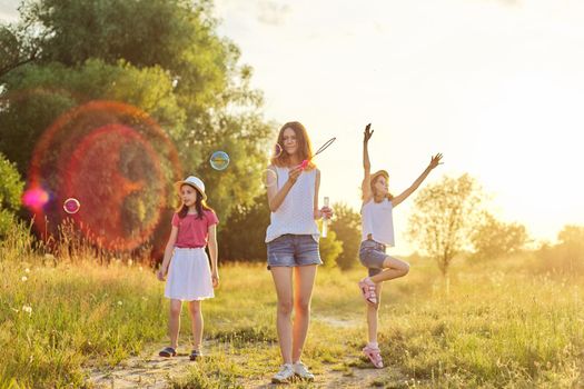 Children three girls playing with soap bubbles, teenager blows younger ones catch bubbles. Beautiful natural landscape, summer meadow background, happy childhood