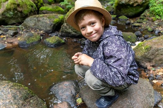 a Caucasian boy in rubber boots, a traveler's hat and a windbreaker is squatting near a forest stream and rocks, looking at the camera and smiling