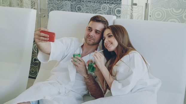 Attractive loving couple is taking selfie with cocktail glasses using smartphone while relaxing in spa salon. They are smiling, talking and posing looking at camera.