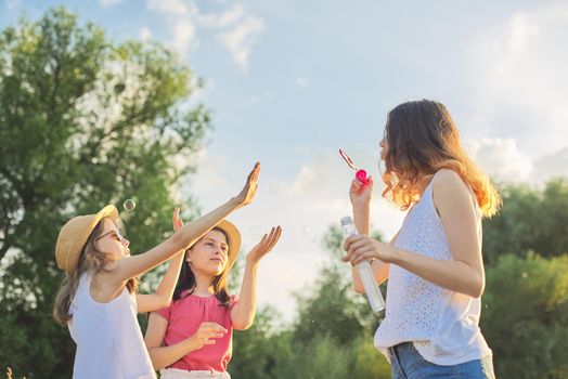 Children three girls playing with soap bubbles, teenager blows younger ones catch bubbles. Beautiful natural landscape, summer meadow background, happy childhood