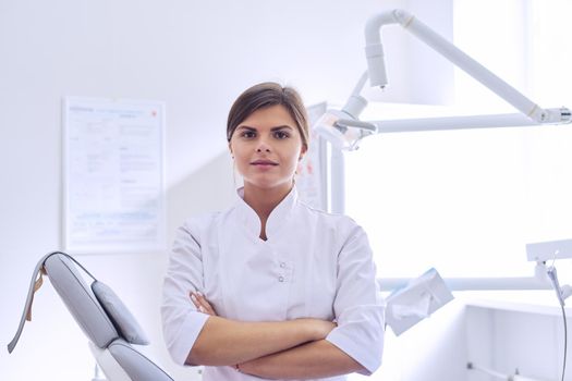 Young positive female dentist doctor with folded arms, dentistry cabinet background