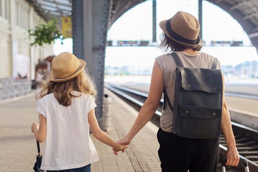 Happy mother and daughter child walking together at railway station with luggage suitcase, back view. Travel, tourism, transportation, family concept