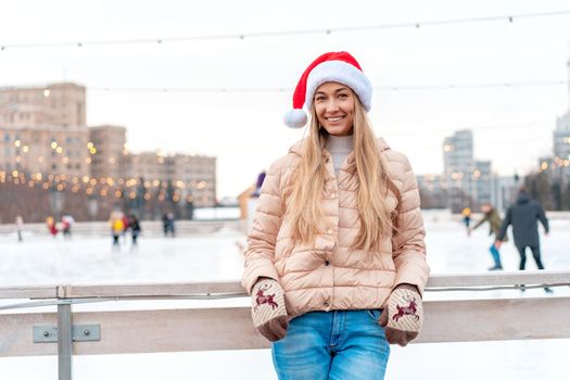 Outdoor portrait young beautiful fashionable woman wearing Santa hat, posing street of European city. Winter Christmas holidays concept. ice skate rink background. Caucasian female winter clothes