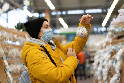 Christmas decoration shopping. Caucasian woman choose new year decor in shopping mall. Medium age female dressed medical mask and yellow winter parka buying Christmas ornament toys indoor store