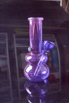 Close-up vertical shot of purple glass bong on a glass surface. Weed smoke device.