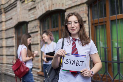 Start. Girl student holds notebook with word start, beginning of classes at school, in college. Brick building and talking students background