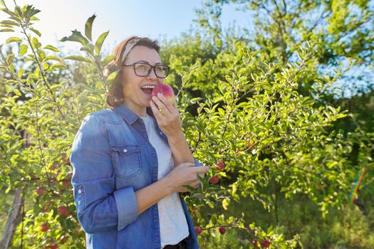 Woman gardener with freshly picked red apple in hand, background is tree with apples. Female eats natural, environmentally friendly apple grown in home garden, copy space