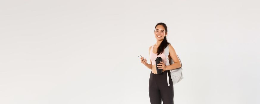 Full length of smiling healthy, slim asian girl going fitness training, female athelte carry backpack with workout equipment and water bottle, using mobile phone sports application, white background.