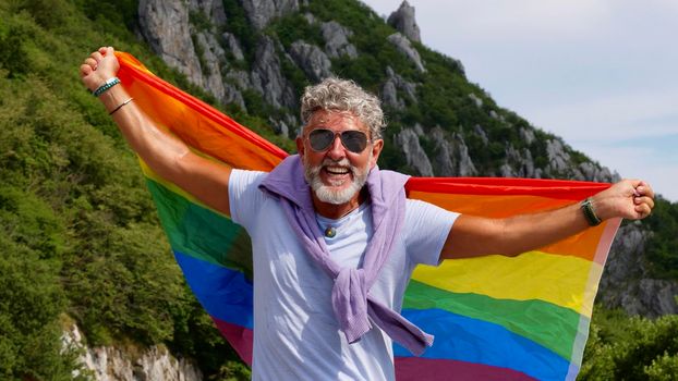 Portrait of a gray-haired senior elderly Caucasian man bisexuality with a beard and sunglasses holding a rainbow LGBTQIA flag on nature. Celebrates Pride Month, Rainbow Flag Day, gay parade