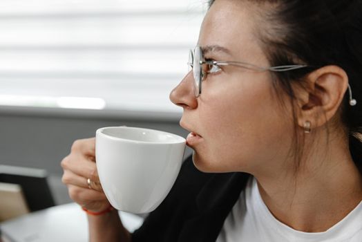 A girl drinks coffee in the office and thinks. A girl is thinking, a break in her work. The employee in the office is not smiling and drinking coffee.