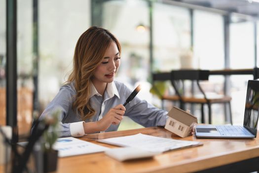 Looking for real estate agency, property insurance, mortgage loan or new house. Woman with magnifying glass over a wooden house at her office.