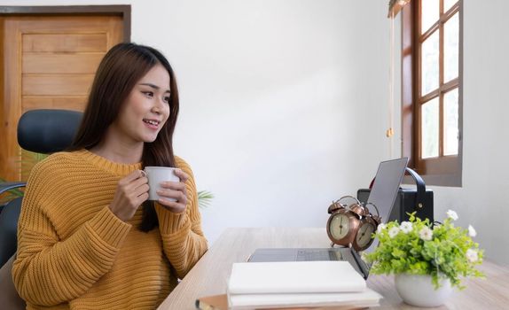 Beautiful young smiling asian woman working on laptop and drinking coffee in living room at home. Asia business woman working document finance and calculator in her home office. Enjoying time at home..